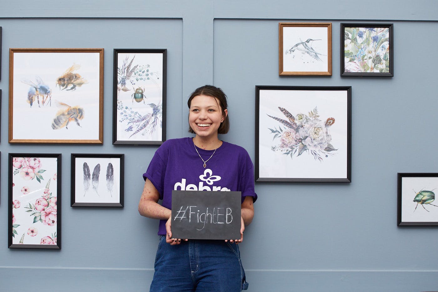 A girl leaning against a lavender coloured wall holding a chalk board that says '#FightEB'. The wall has paintings hanging on it.