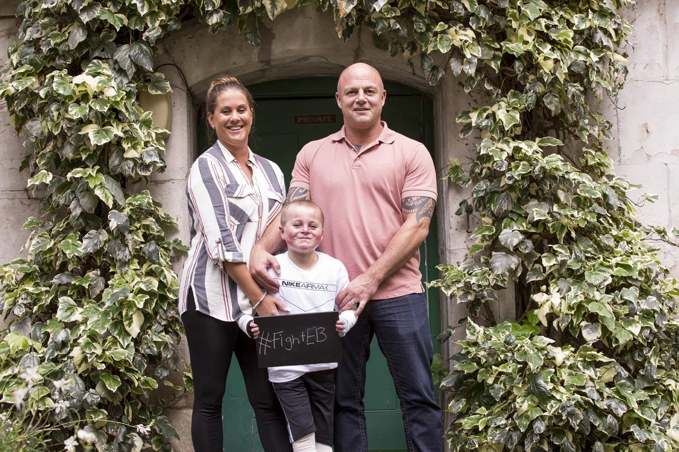 A family of mother, father and son. Son is in the middle holding a chalk board saying '#FightEB'. Standing in front of a door with ivy growing all around.