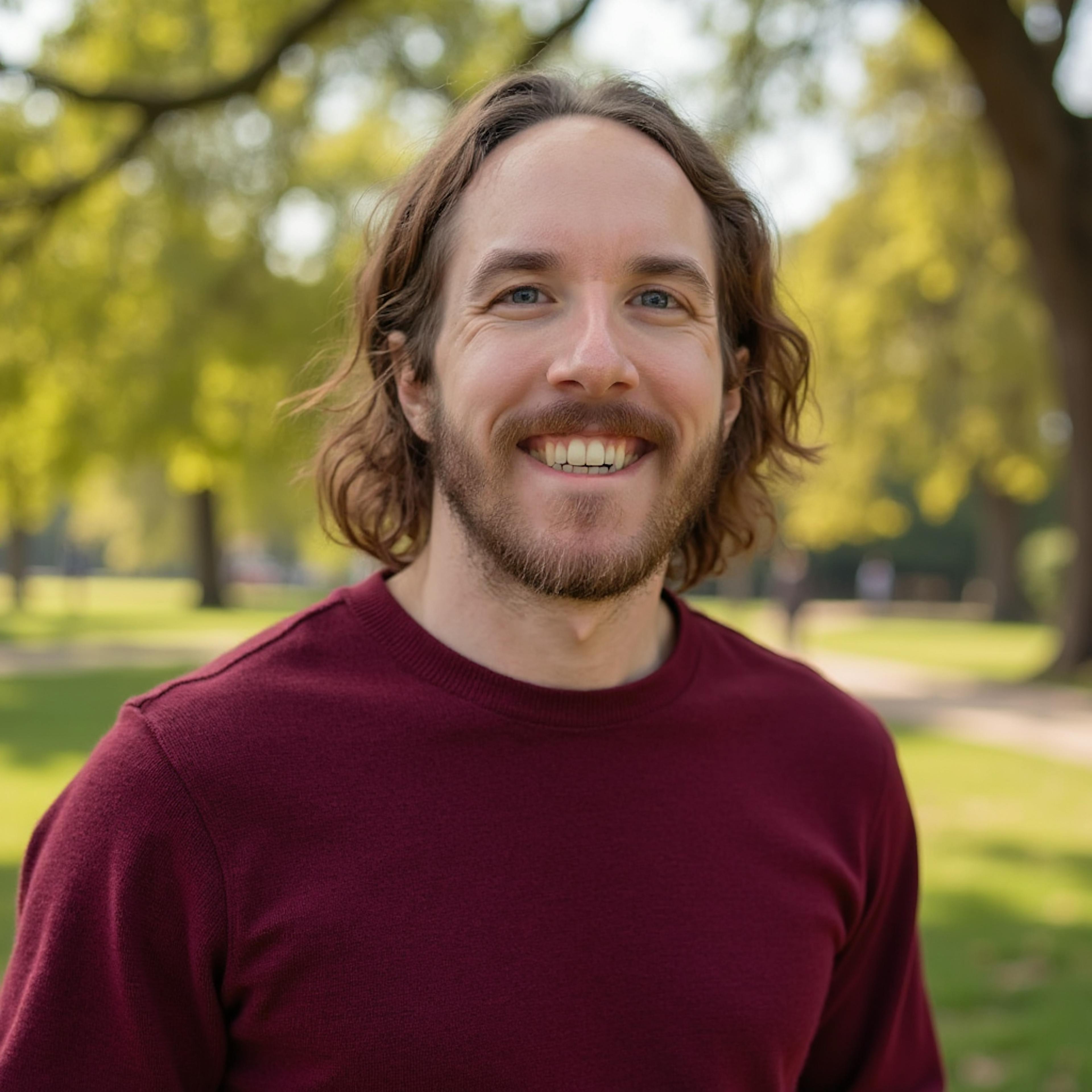 Photo of a man with brown, shoulder-length wavy hair and a beard smiles at the camera. He is wearing a maroon sweater and is outdoors, with a park setting and trees in the background.