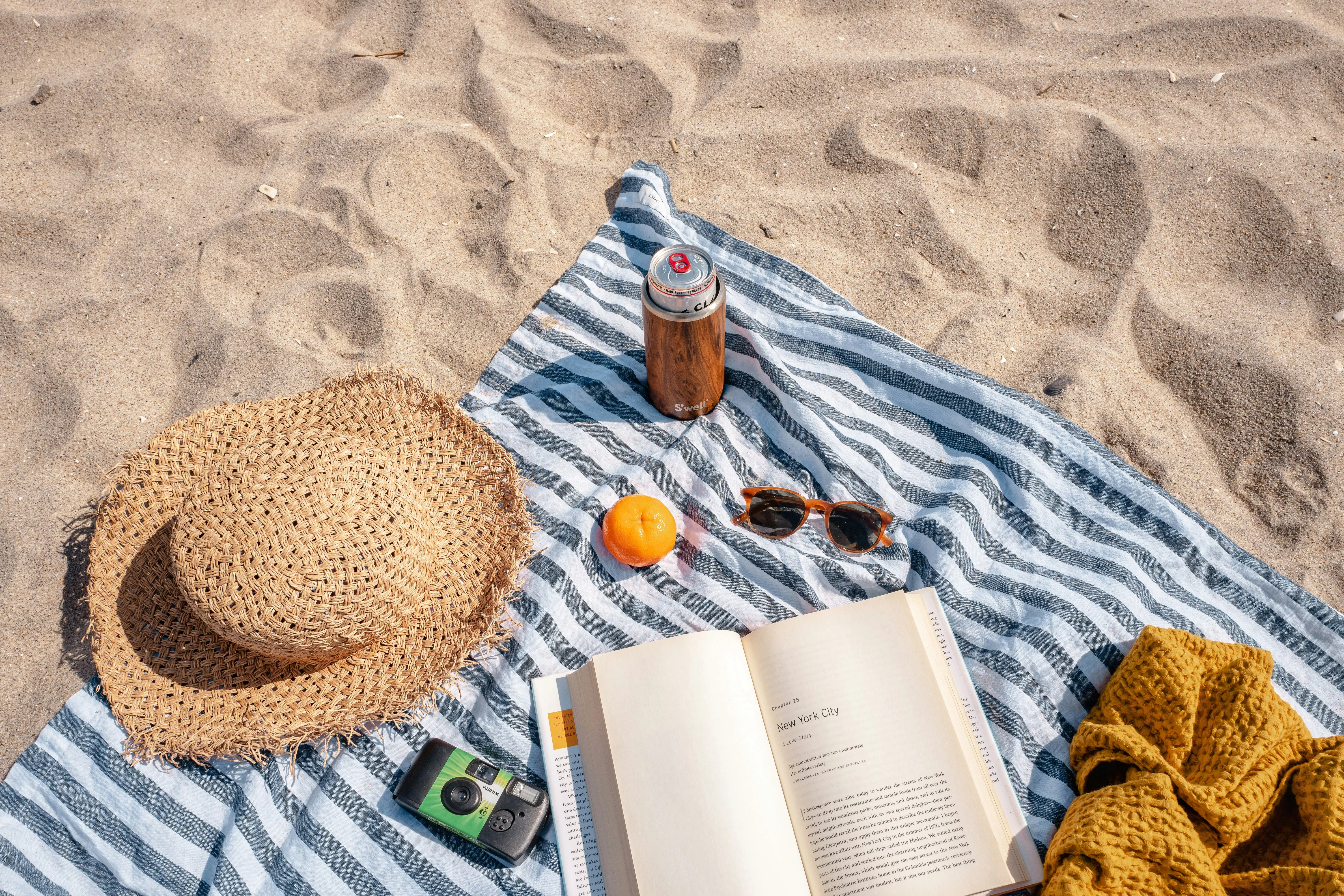 toalha de praia na areia com chapéu, bebidas e livro