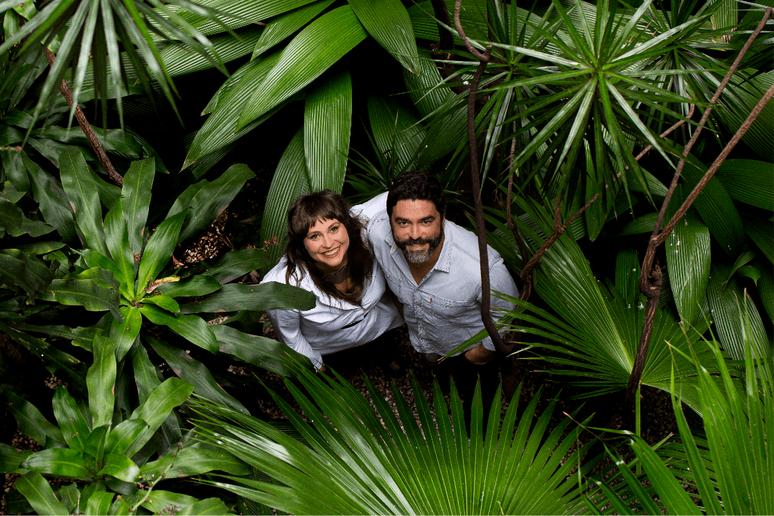 Visão superior de Carol Ayres e Bruno Gomes, que olham para cima sorrido, enquanto estão abraçados lado-a-lado no meio de uma diversidade de plantas com folhas verdes-vivo.