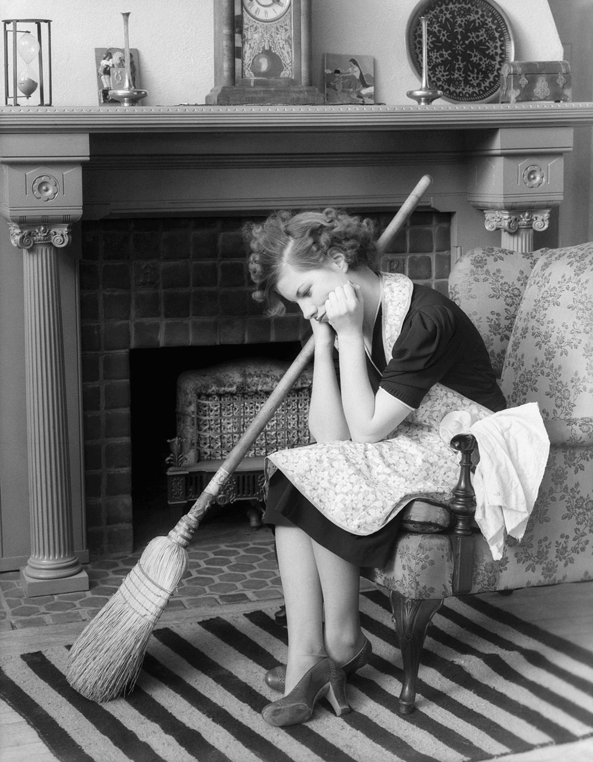 Black and white photo of an unhappy woman sitting on a chair head down in front of fireplace. She is wearing an apron and has a broom at her side.