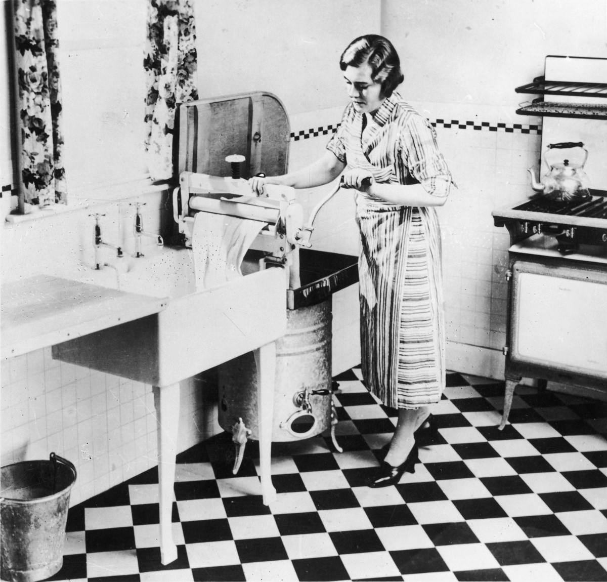 black and wife picture of a wife running some washing through a mangle in her kitchen.