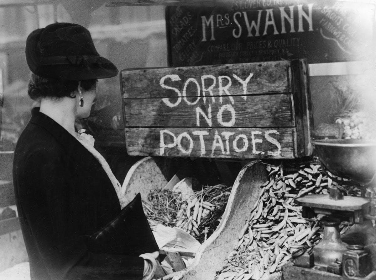 A black and white photograph of a grocery store, where a well-dressed woman looks at empty shelves, with a make-shift sign saying Sorry No Potatoes