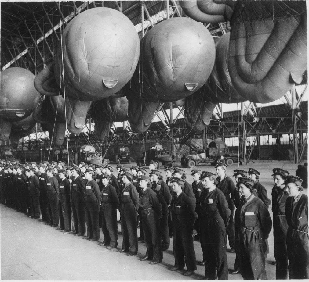 Black and white photo of a W.A.A.F. operators in a hangar. They are all standing still in uniform, with their hands behind their back and in a row as they are reporting for inspection at the end of a day of training. Behind them, the ballons have been tucked away and can be seen floating in the hangar.