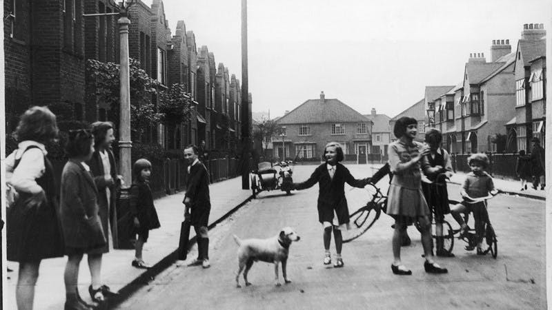 A black-and-white photograph of a group of children playing in a quiet street. There are several bikes, one of them is skipping with a rope, and there is a small dog looking on.