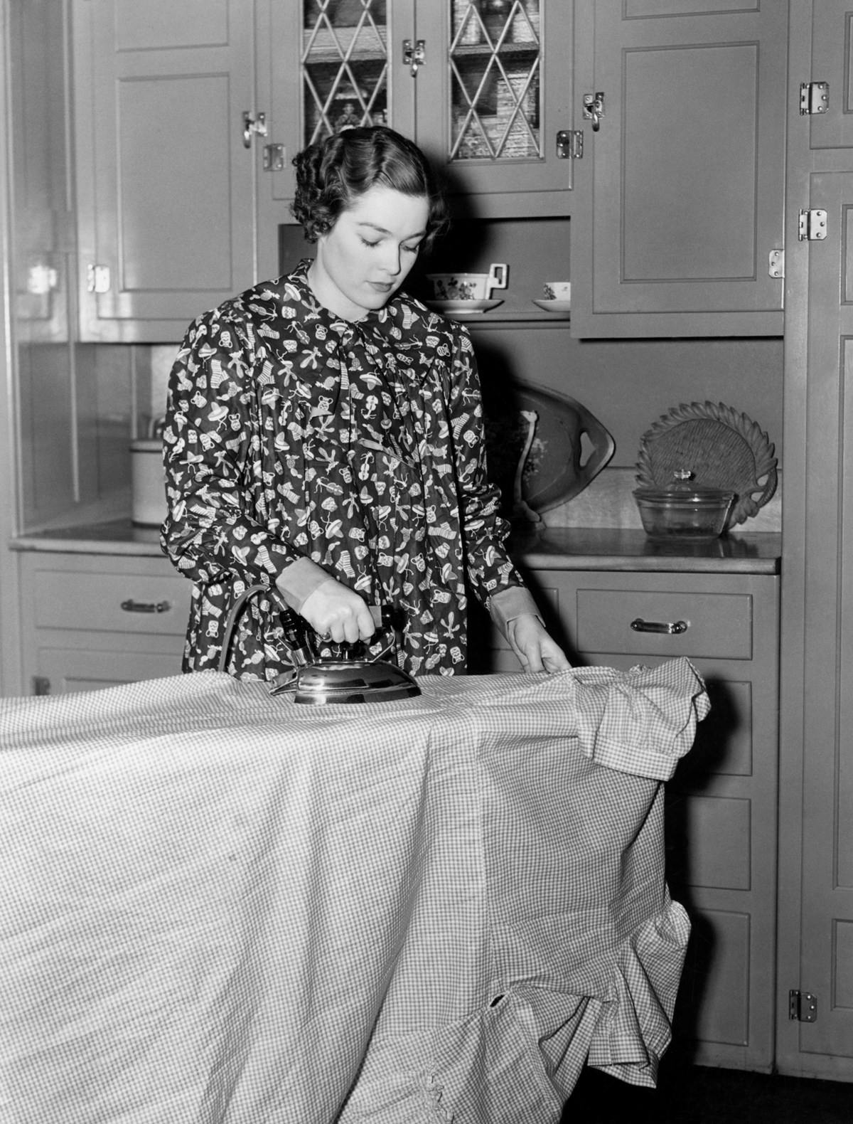 Black and white photo of a housewife using an electric iron on a checkered dress or apron. The woman is standing in front of cupboards of kitchenware. She is wearing a printed smock apron with little sombreros, cacti, scarves and musical instruments.