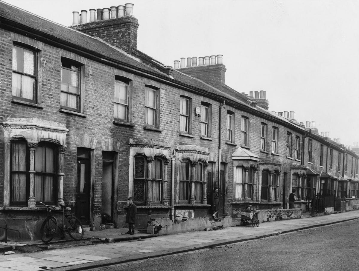 Black and white photograph of a row of run-down terraced houses. A bike rests against a window, and a small boy looks on.