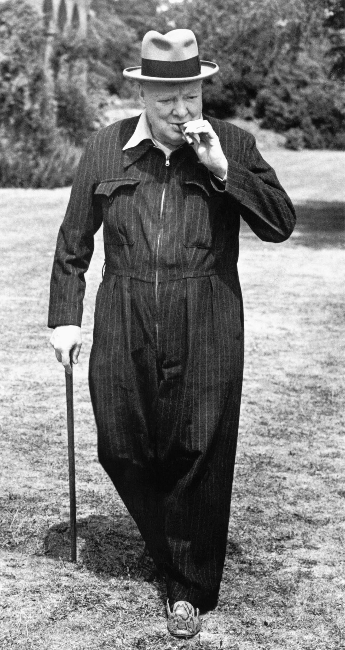 A black and white photograph of Winston Churchill. He is wearing a siren suit, and hat, and is leaning on a cane in his right hand, and is smoking a cigar held in his left.