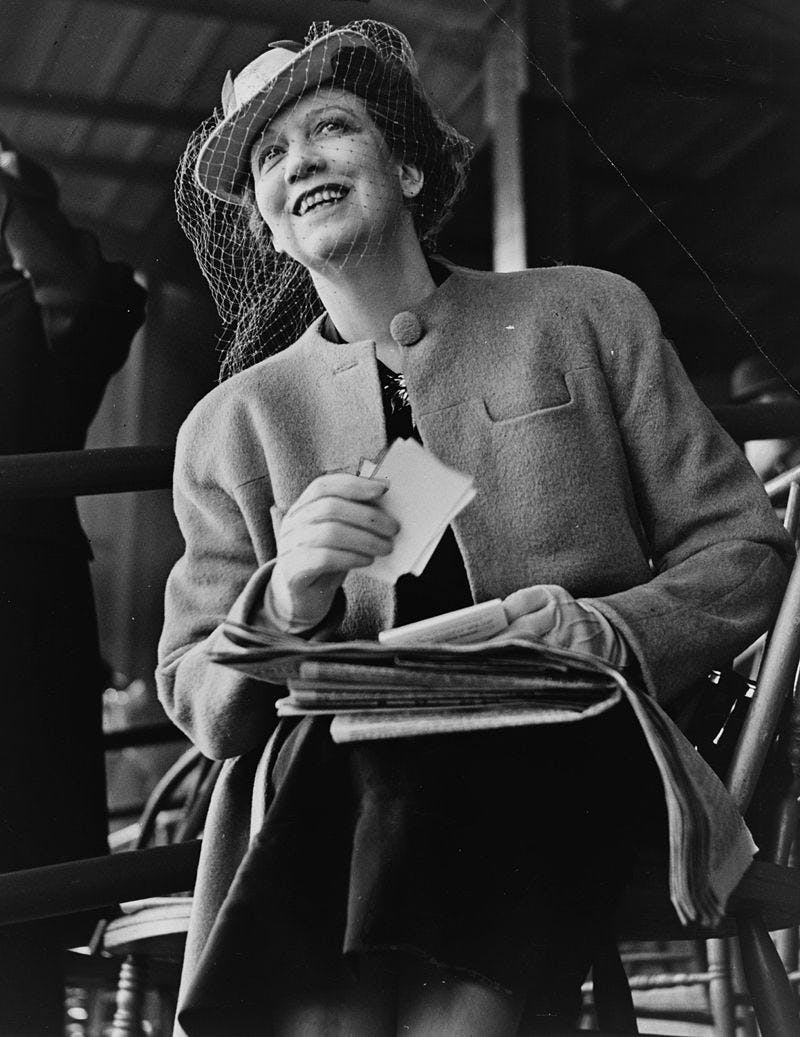 A black and white photograph of Elizabeth Arden sitting with a newspaper on her lap and white slips of paper in her hand, wearing a stylish hat and jacket.