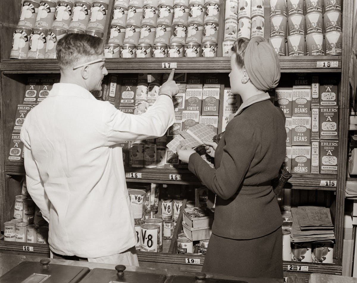 A black and white photograph of shelves of tinned goods in a grocery store. A customer holds her ration book, while a grocer points at a price.