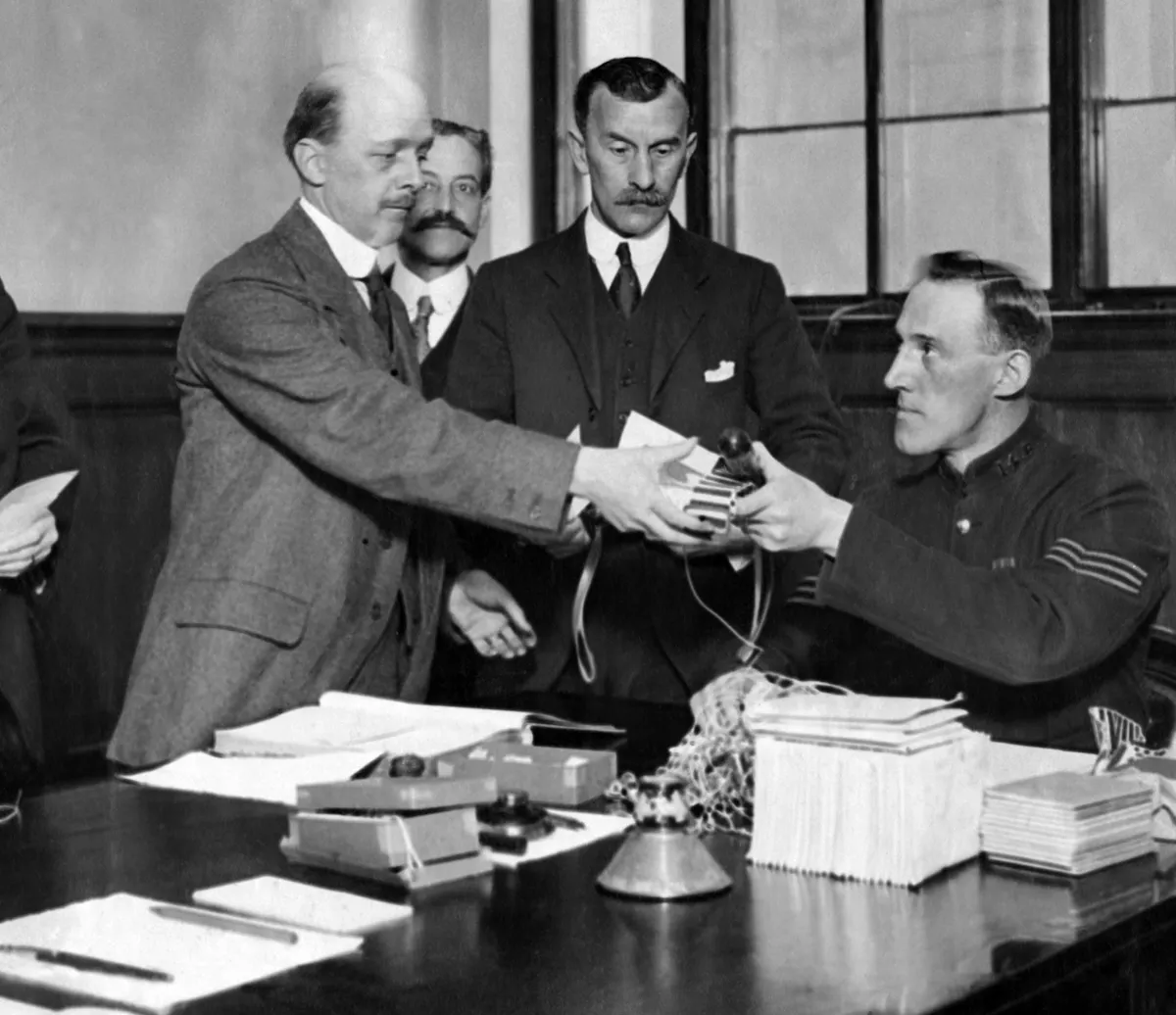Black and white photo showing a man seated at a desk in uniform, handing a man standing some papers.