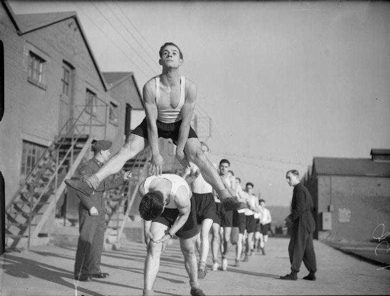 Black and white photo of soldiers from the Hampshire regiment during physical training at Oakridge farm in Bassington. The men training are performing leapfrog exercises in line. They are all wearing black shorts and white vests. A superior and another unidentified man are watching them.