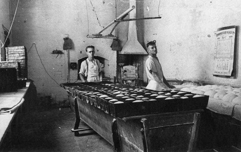 Black and white picture of the interior of Dumpleton's bakery in 1939. There are two bakers wearing apron looking at the objectives. You can also see a bench of loaves rising on the right side.