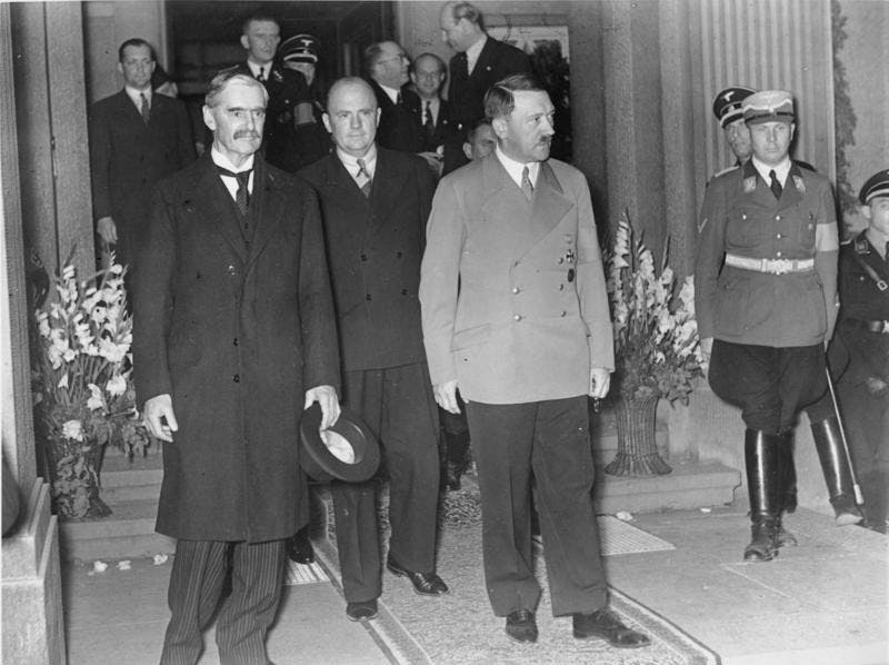 A black-and-white photograph of Neville Chamberlain and Adolf Hitler walking out of a grand entrance-way. In the background, other gentlemen in suits converse.