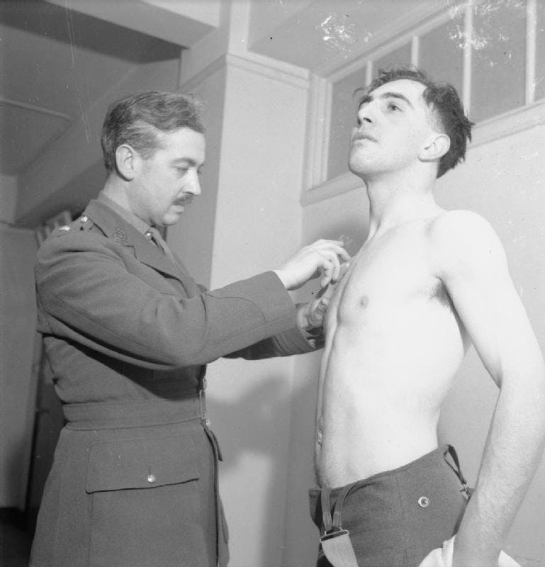Black and white photo of a young man, Bill Jones, passing his medical examination to enter the army. He is standing without his shirt on the right. The army doctor is on the left, in uniform and wearing a mustache.