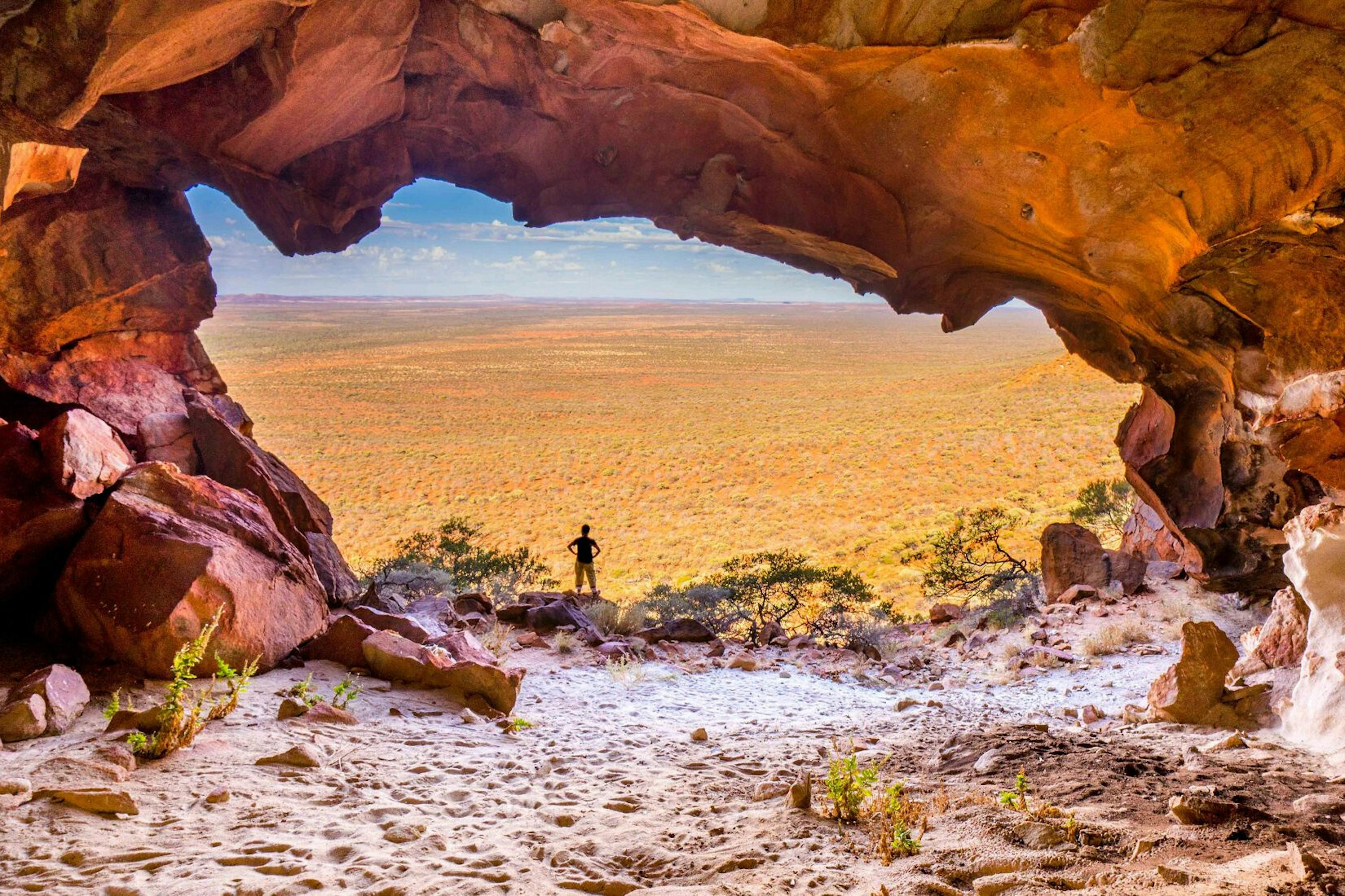 A photo of a person, framed by what appears to be a cave, looking out onto the red plains of Australia. Photo credit: John Spies via The Nature Conservancy