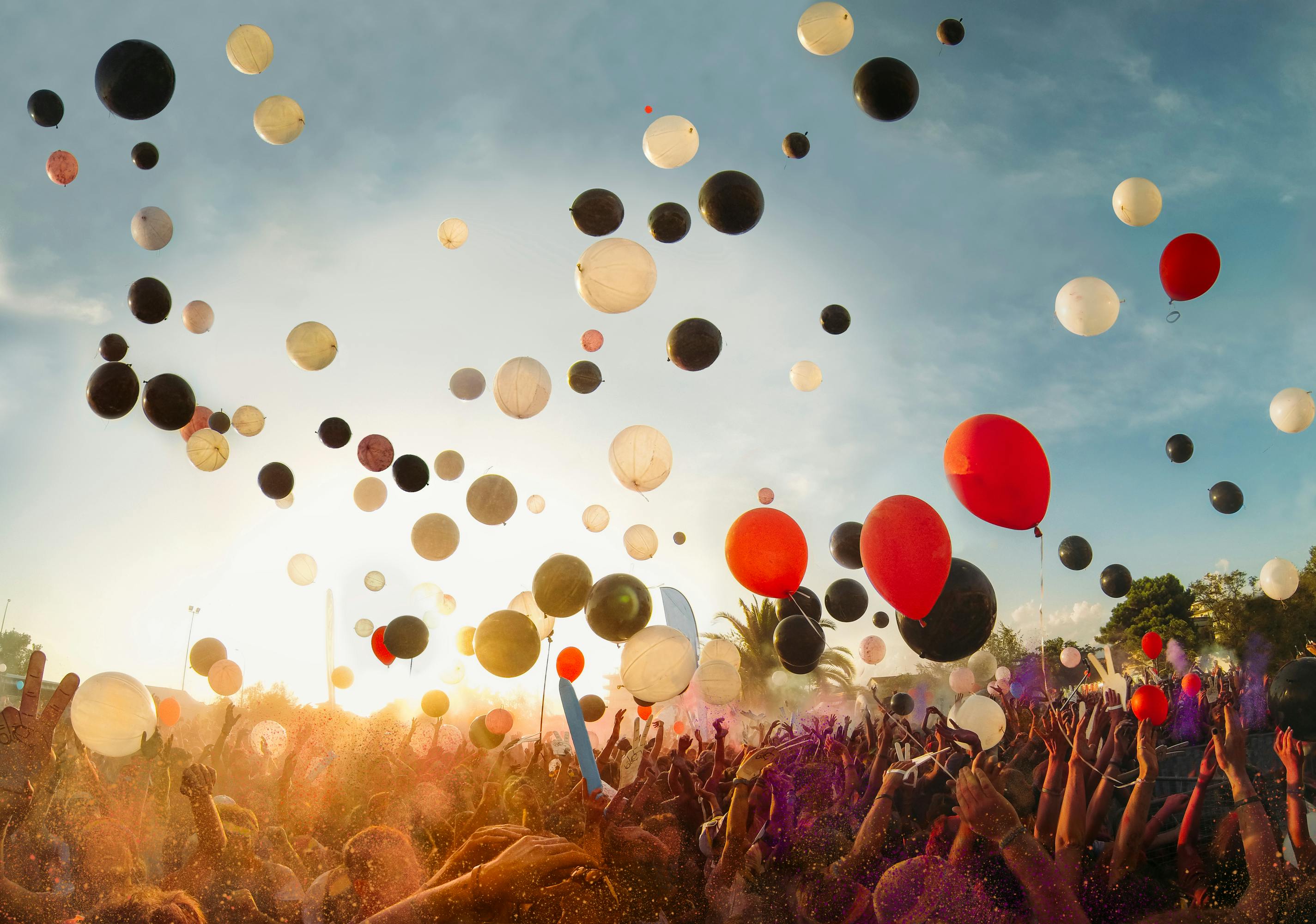 A crowd of festival revellers waving their arms in the air while balloons fall from the sky, the sun glistening on the horizon.