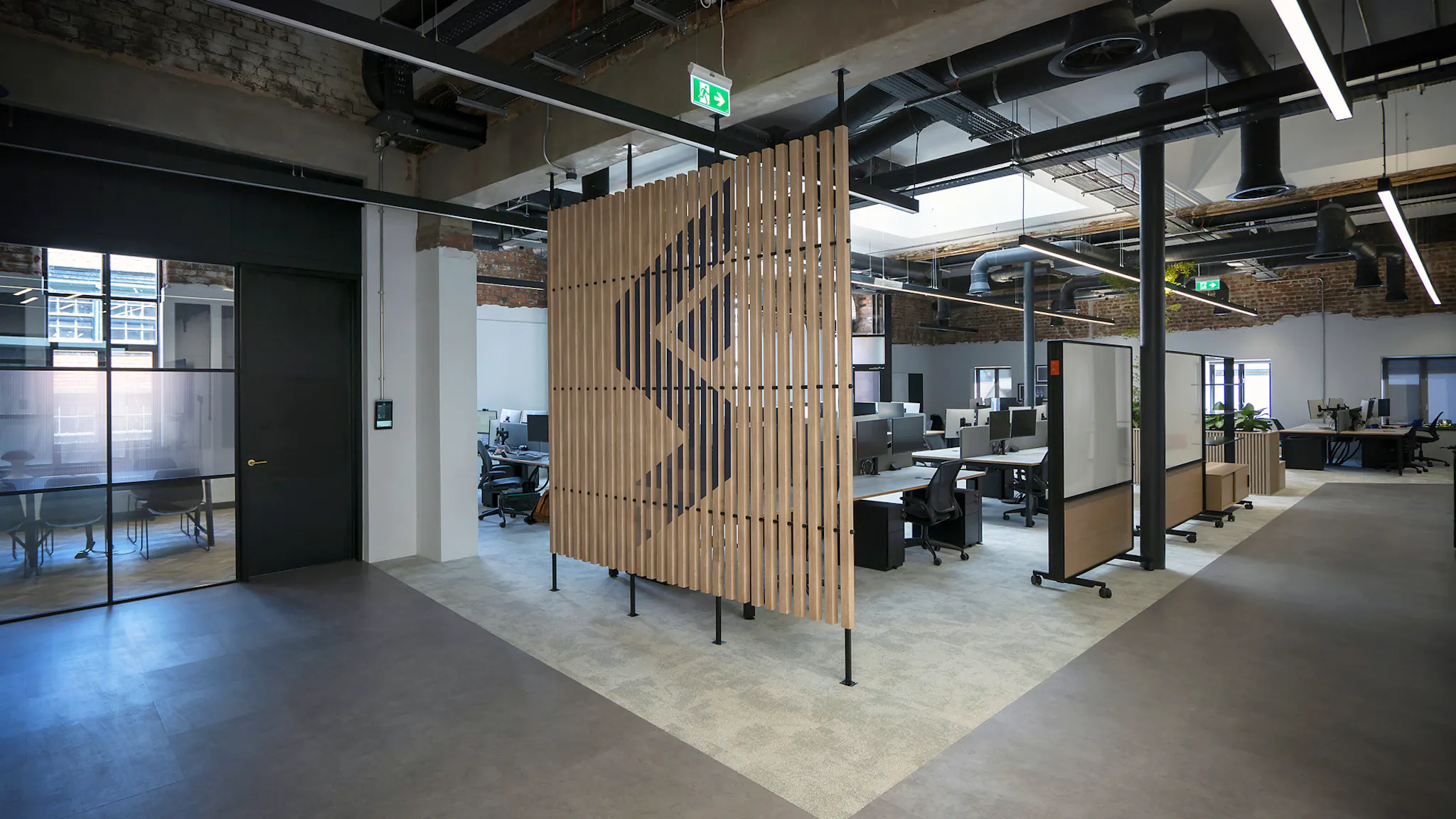 A game studio setting with meeting room visible to the left and a clutch of desks behind a large decorative screen of wooden slats which displays the Firesprite logo icon in black.  