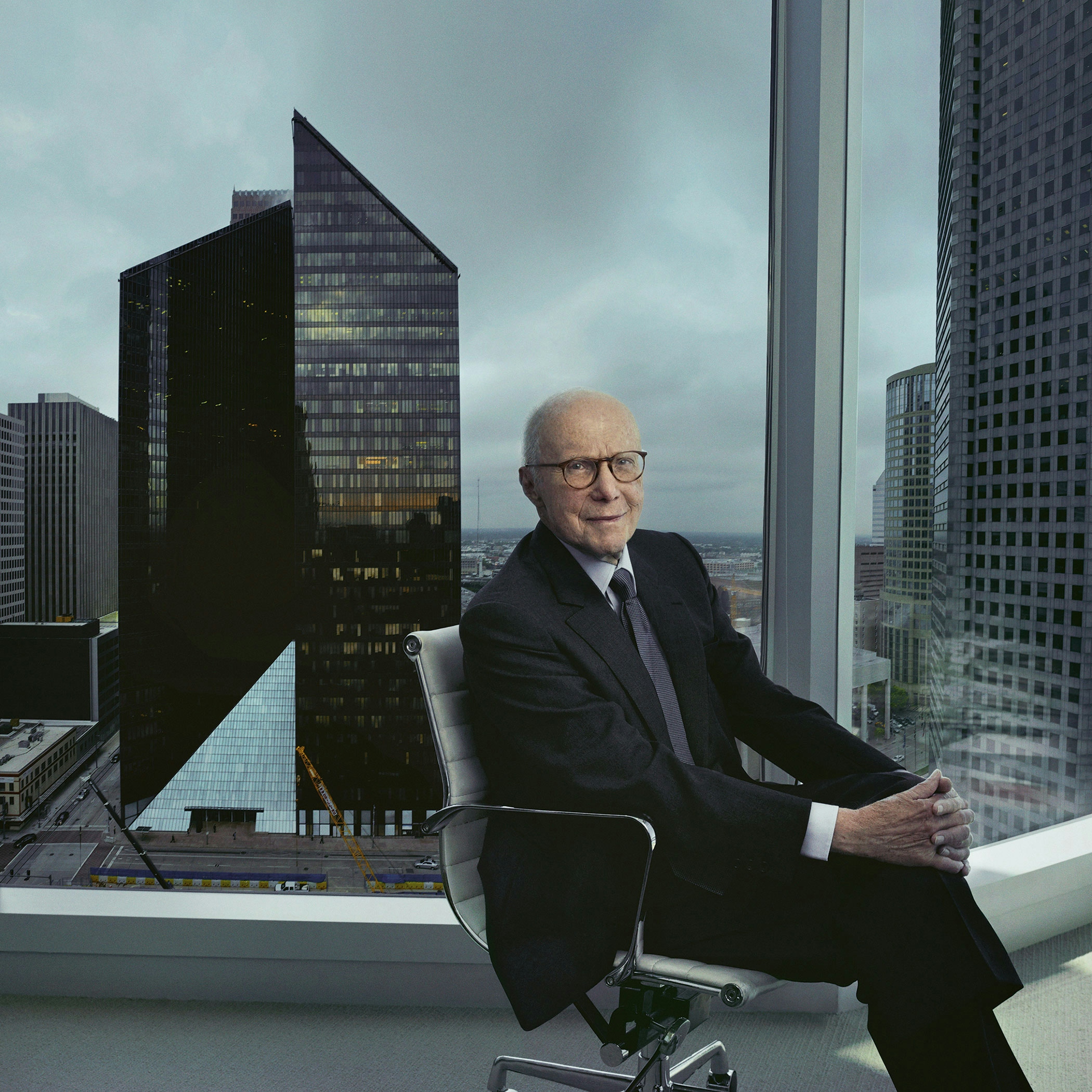 Gerald D. Hines in a business suit sitting in a chair in an office with a city below him in the background.