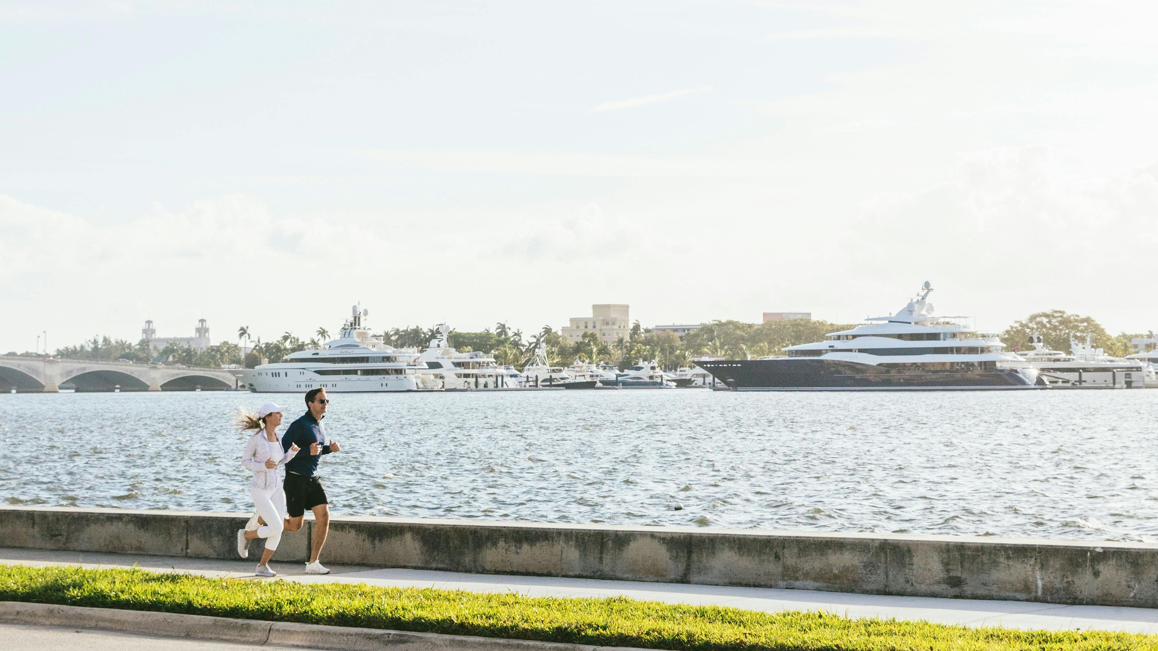 A man and woman in fitness attire running on a sidewalk with a yacht marina in the background.