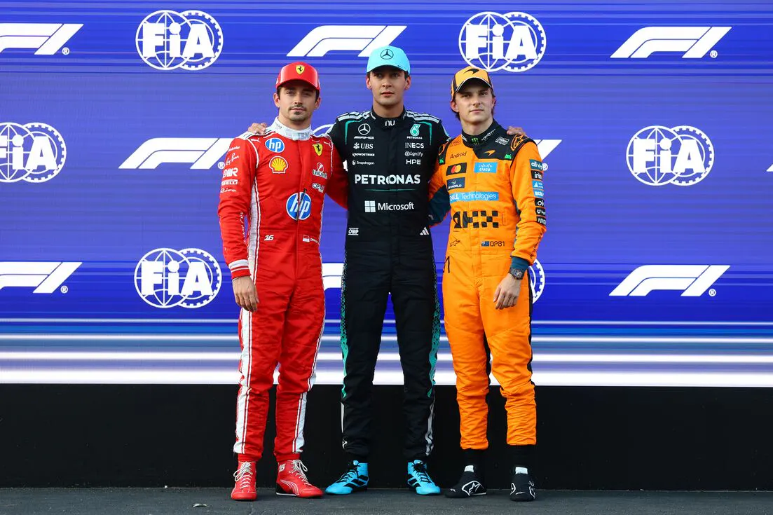 Three F1 racers stand embraced posing in front of a blue F1 backdrop