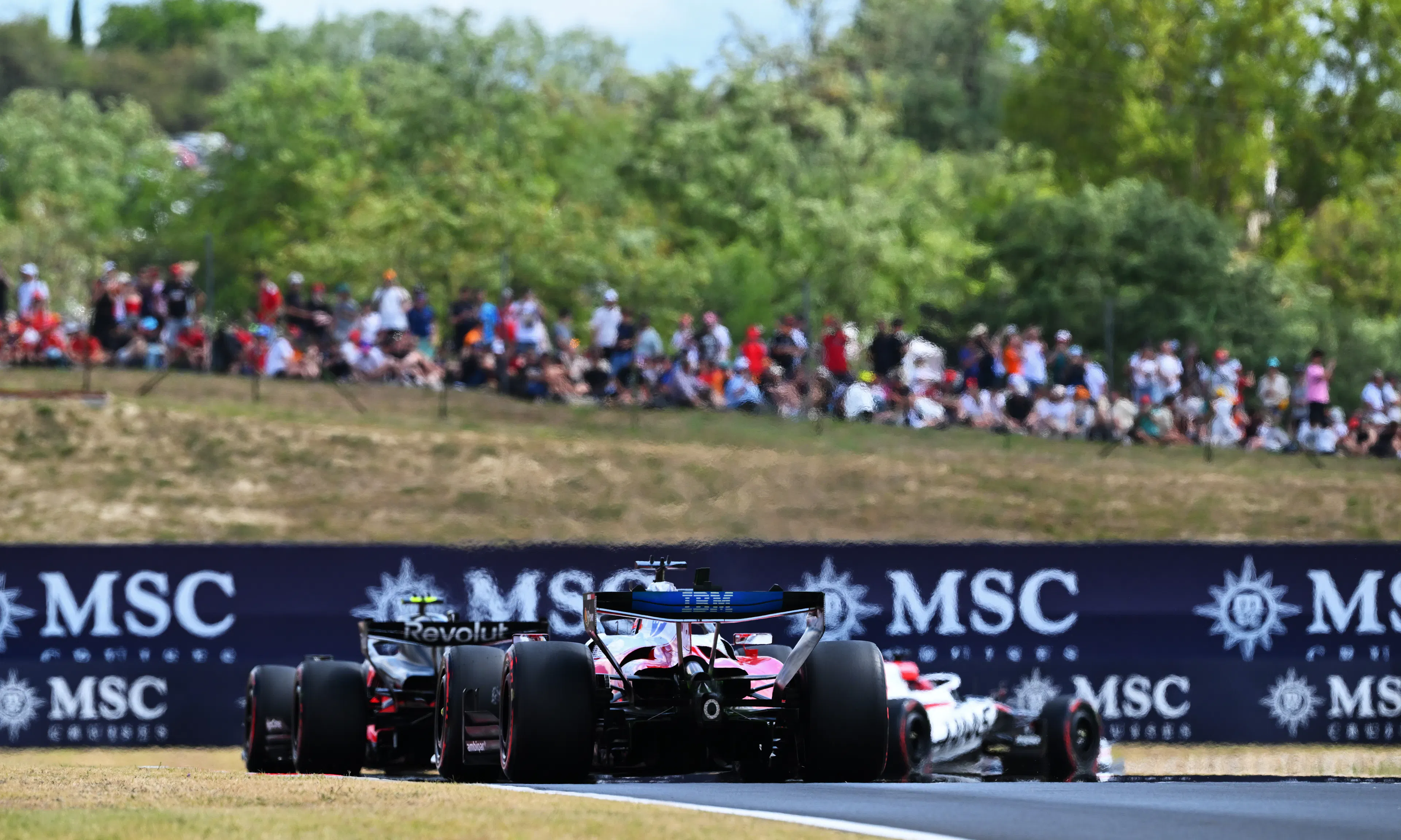 F1 cars taking a turn with spectators on a small hill above watching