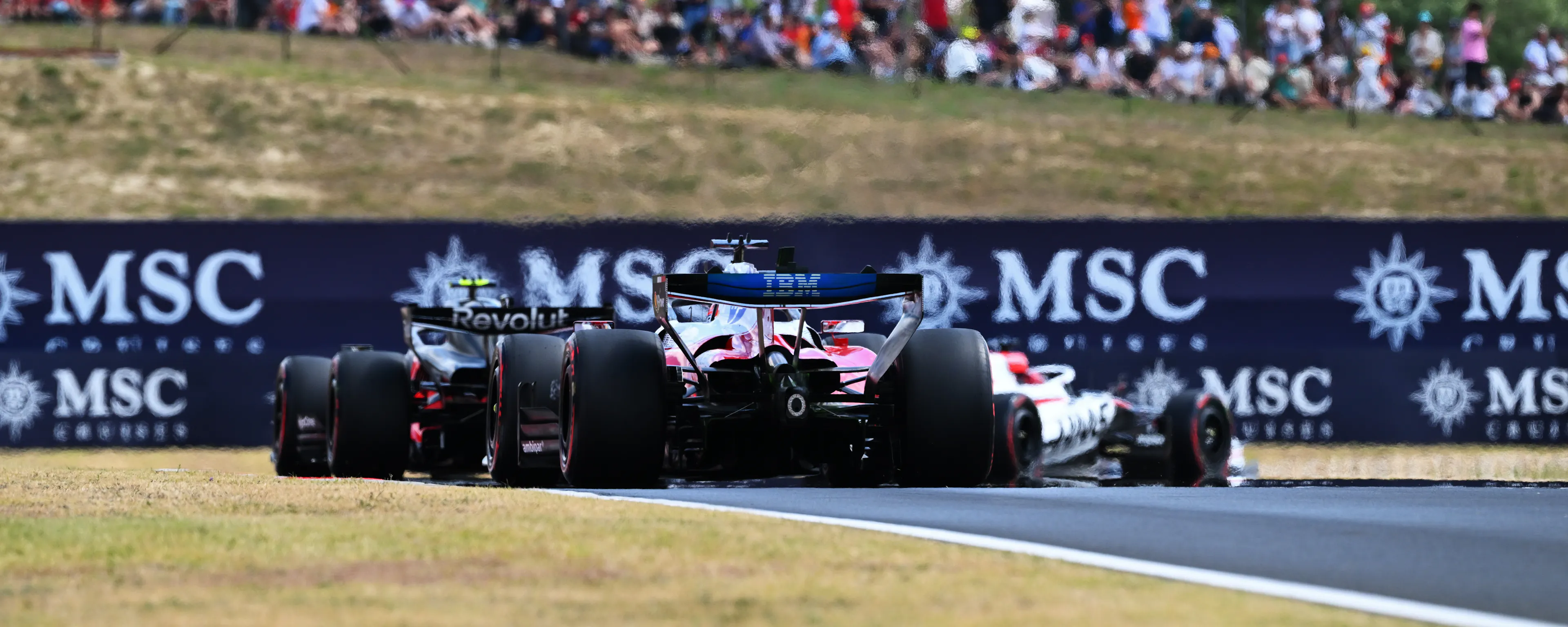 F1 cars taking a turn with spectators on a small hill above watching