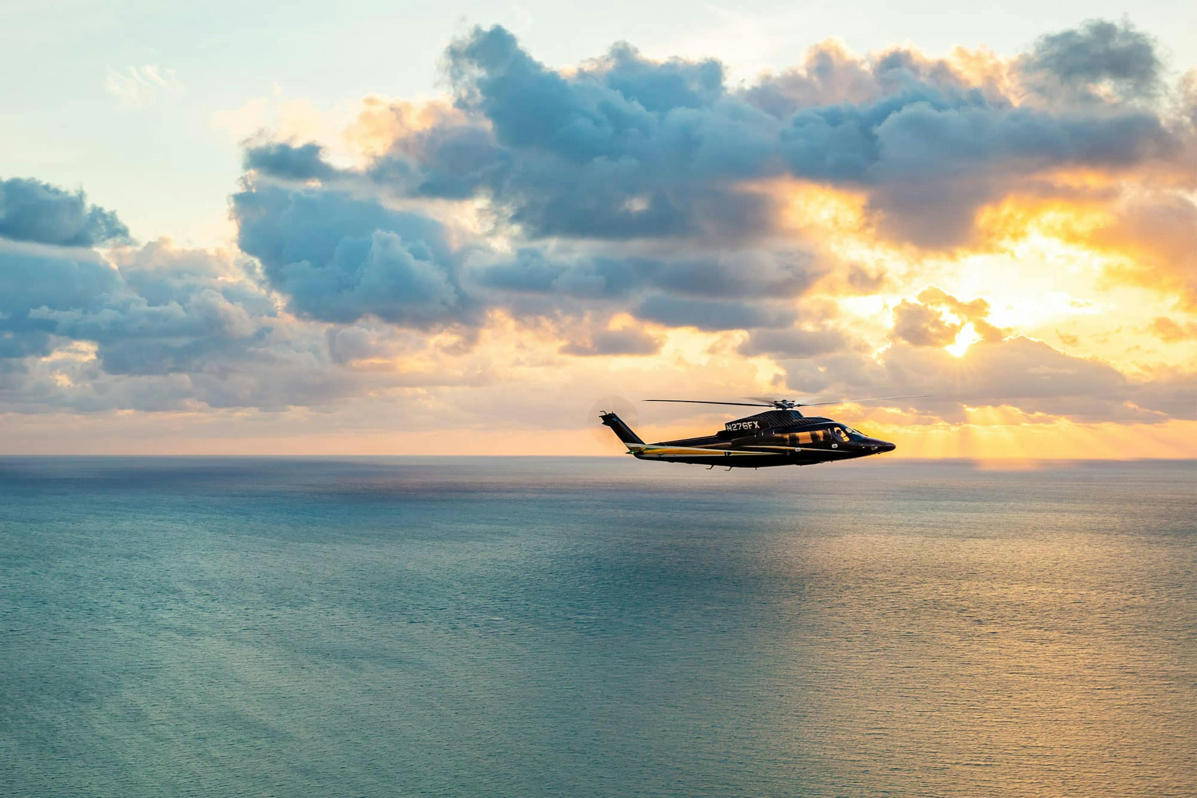 An S76 flies over slightly choppy waves of water with sun beams peaking through the clouds. 