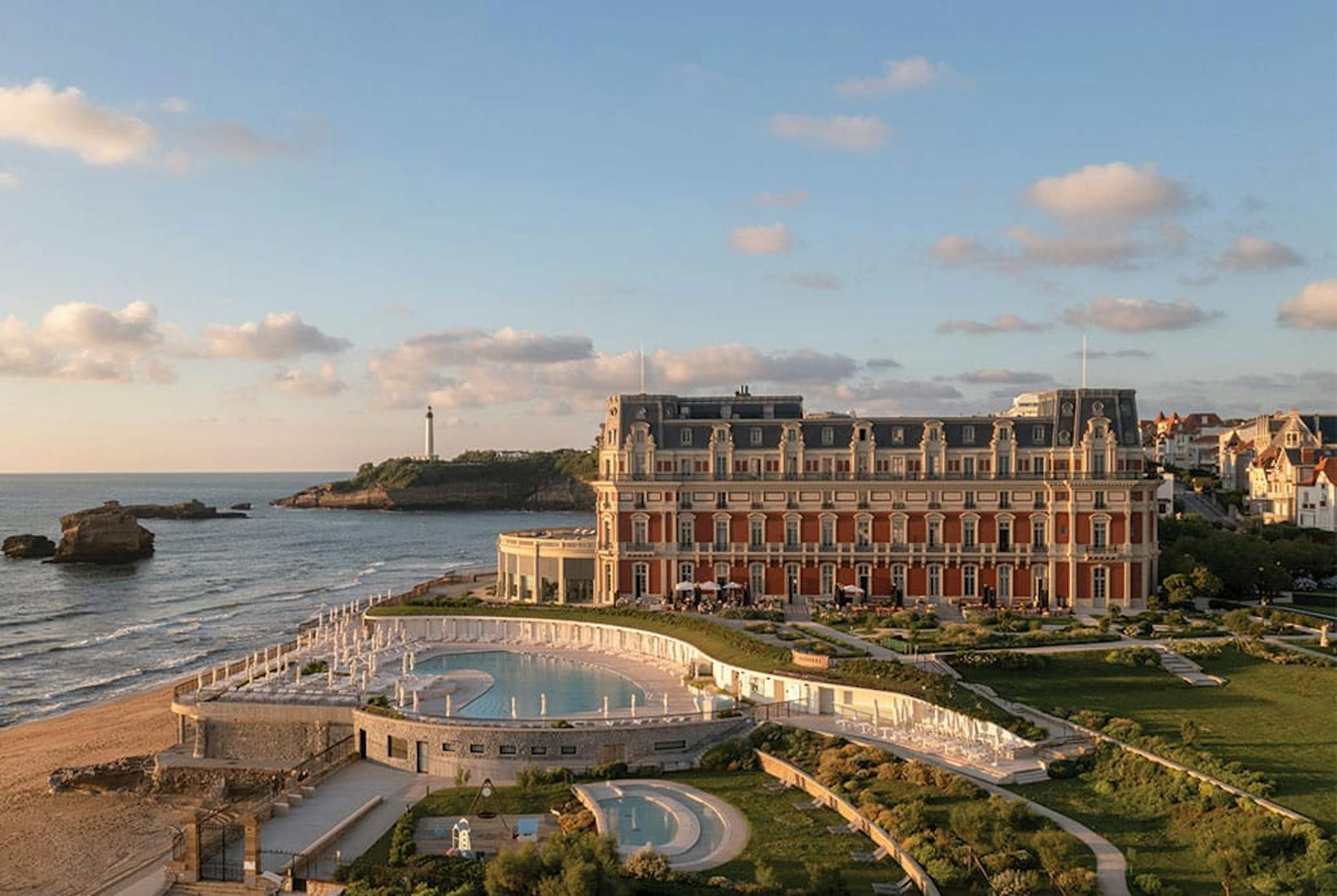 Grand hotel with complext pool and deck sits on the water with lighthouse in the distance