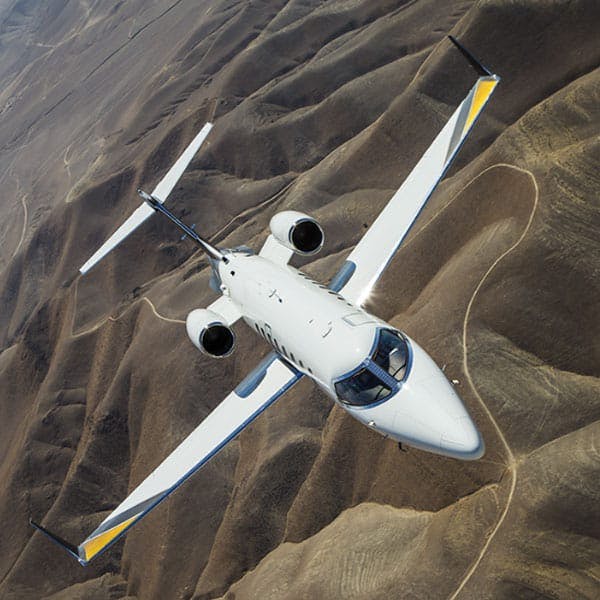 A small private jet flies over rolling hills with a road carved through
