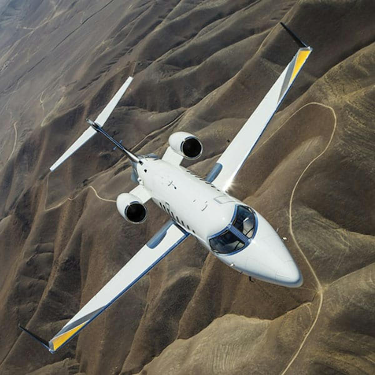 A small private jet flies over rolling hills with a road carved through