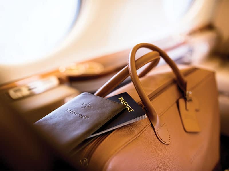 close up of a passport and passport case sitting on brown leather bag closed next to a window of an aircraft