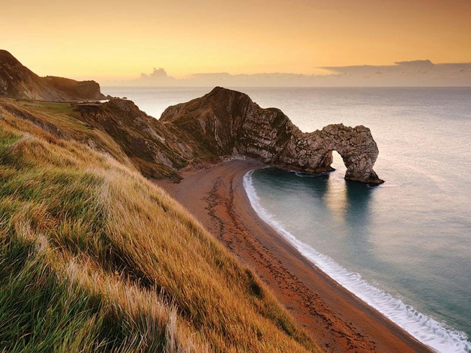 Rocky archway into sea from a beach, viewpoint up on a grassy hill