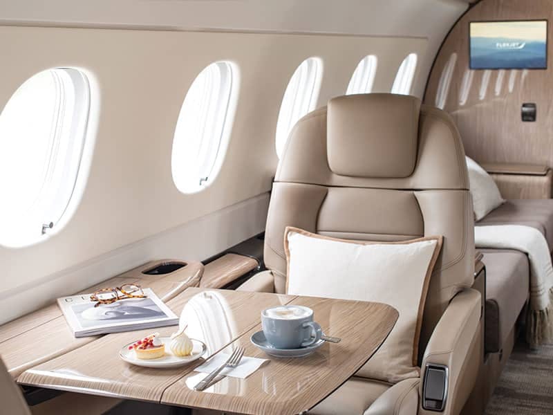 a magazine, some food on a plate, and a cup of latte or coffee sits on a table extended from a window in a plane