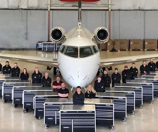 several mechanics posing in front of a jet with their black and chrome tool boxes in front of them all