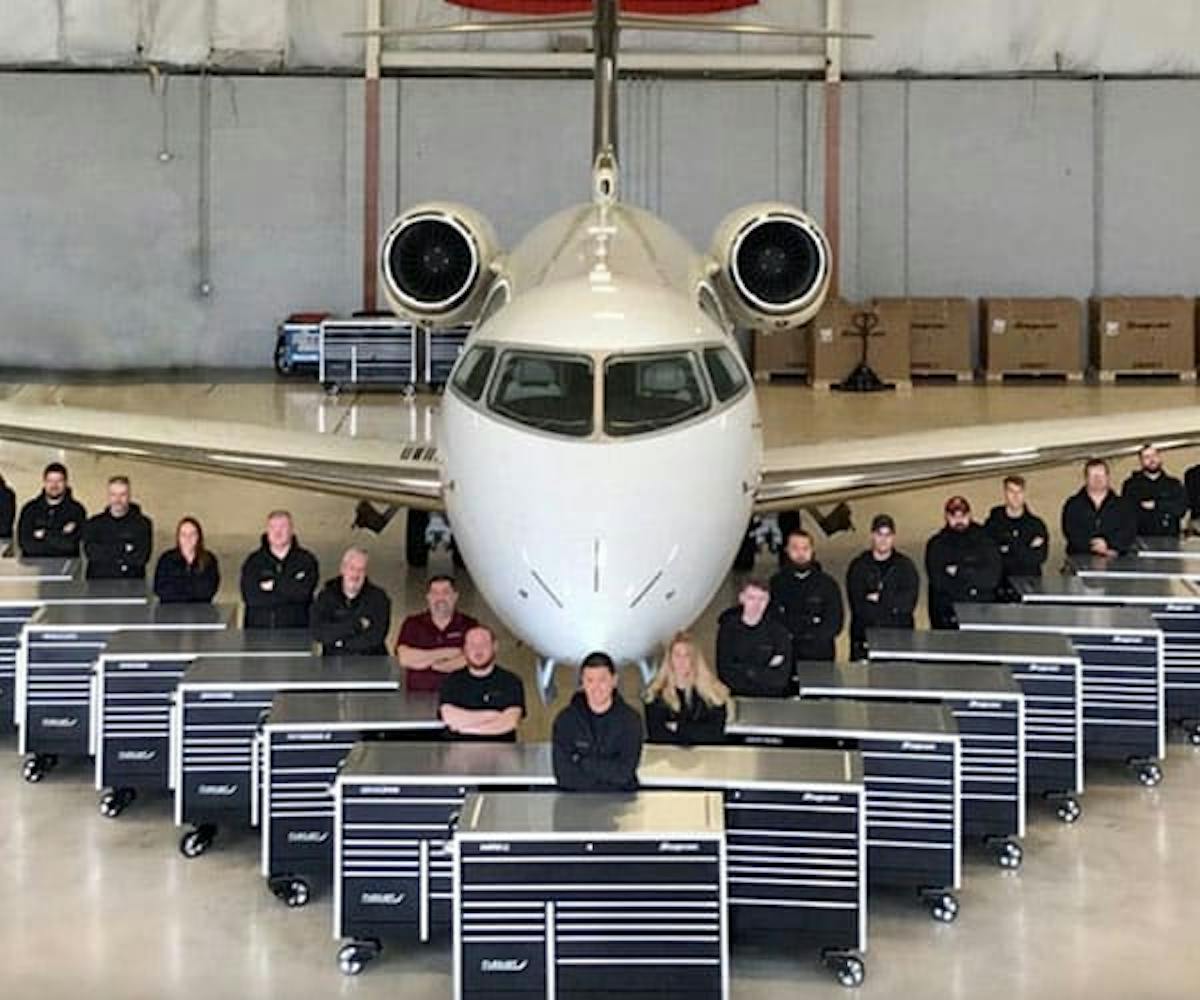 several mechanics posing in front of a jet with their black and chrome tool boxes in front of them all
