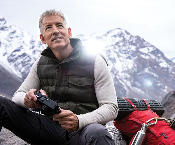 An older man in an outdoor vest and with a backpacking pack, waterbottle, and bedroll, sits on part of a mountain admiring the scenery with a camera in hand