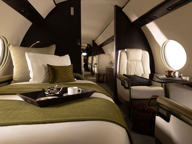 A tray with a coffee cup and kettle sit on a nicely made day bed sofa combo across from a luxurious seat and table on a private jet