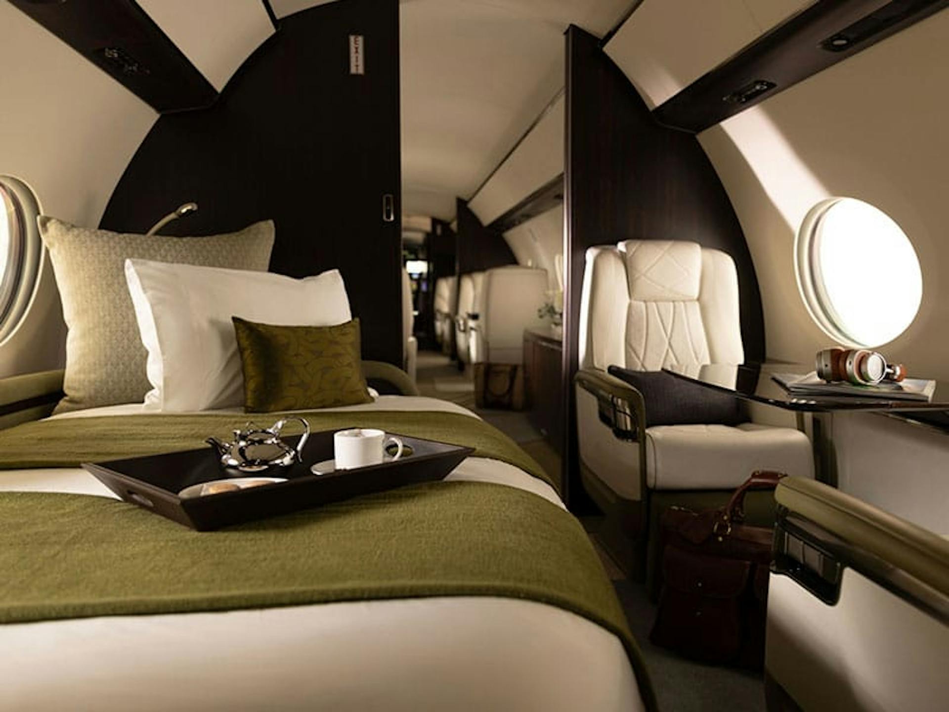 A tray with a coffee cup and kettle sit on a nicely made day bed sofa combo across from a luxurious seat and table on a private jet