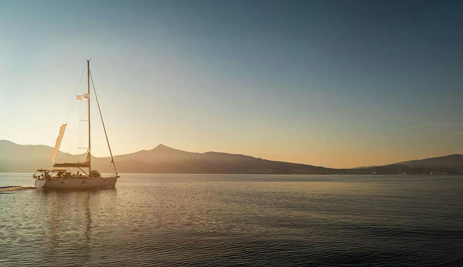 A solo sailboat on water with the sun down low behind mountain ridges