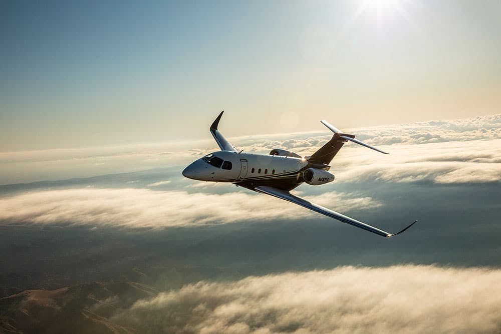 A praetor 500 soars above the clouds with a slight glimpse of the sun at the top of the photo reflecting down on the aircraft