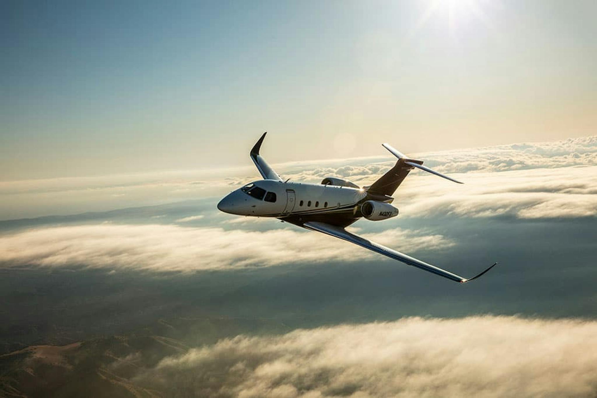 A praetor 500 soars above the clouds with a slight glimpse of the sun at the top of the photo reflecting down on the aircraft