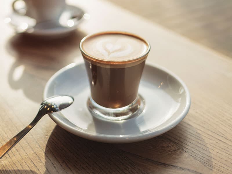 latte in a glass filled to the brim with a heart design on the foam, spoon resting in the dish under it