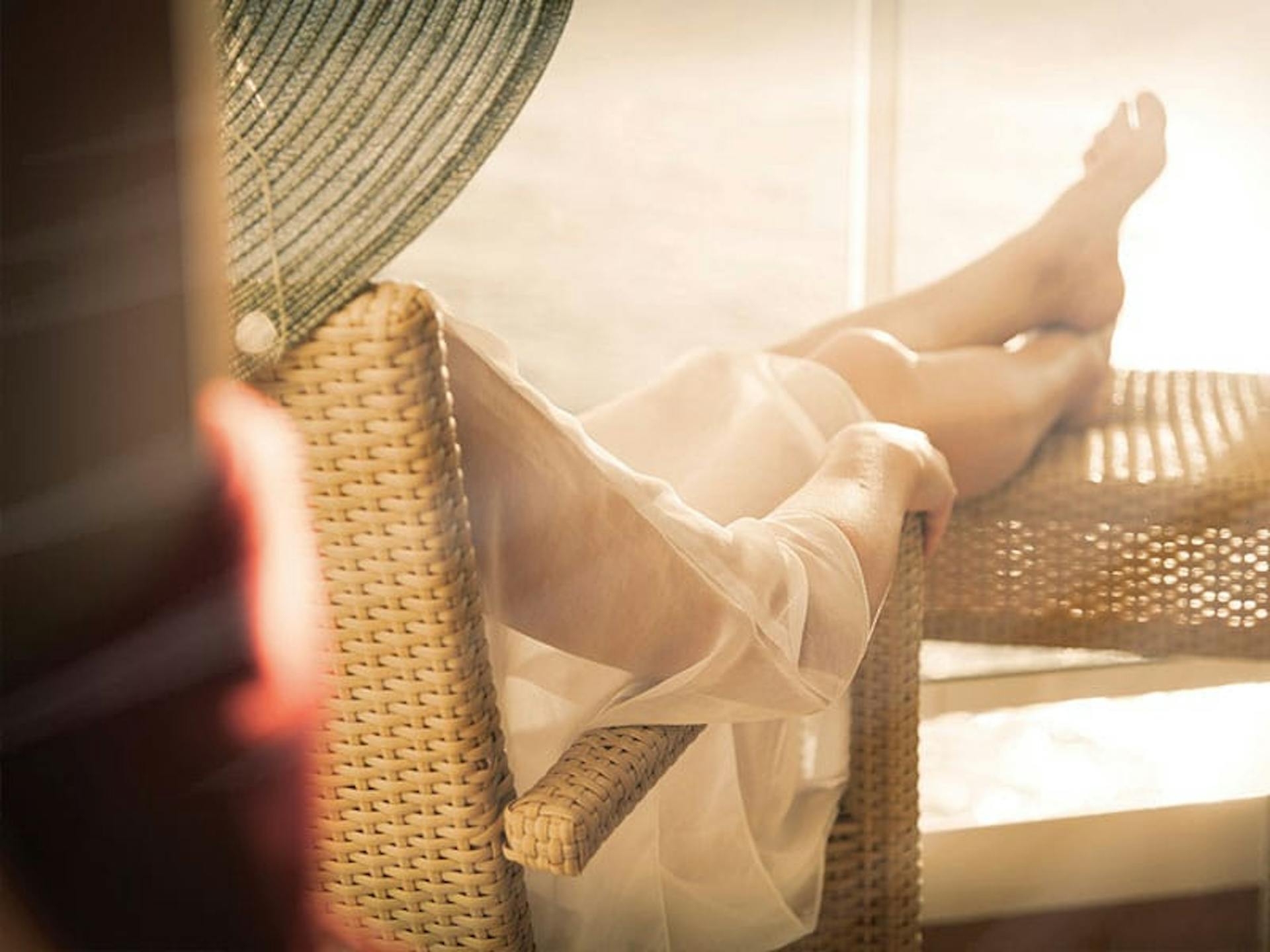 Someone with their legs kicked up on an outdoor table, part of their sun hat showing as well, as they face the direction of the sunlight on a porch