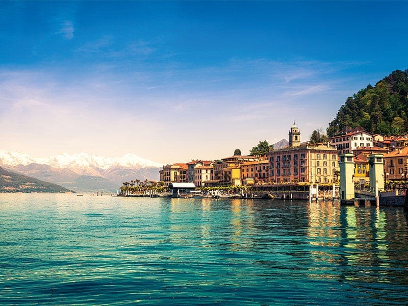 blue rippling waters at the foot of a town with light brown old buildings and in the distance is white capped mountains