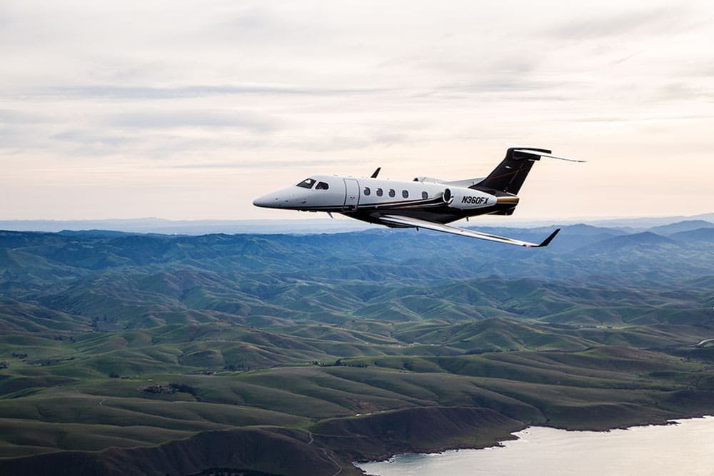 A Phenom 300 flies over rocky rolling hills and a bay of water