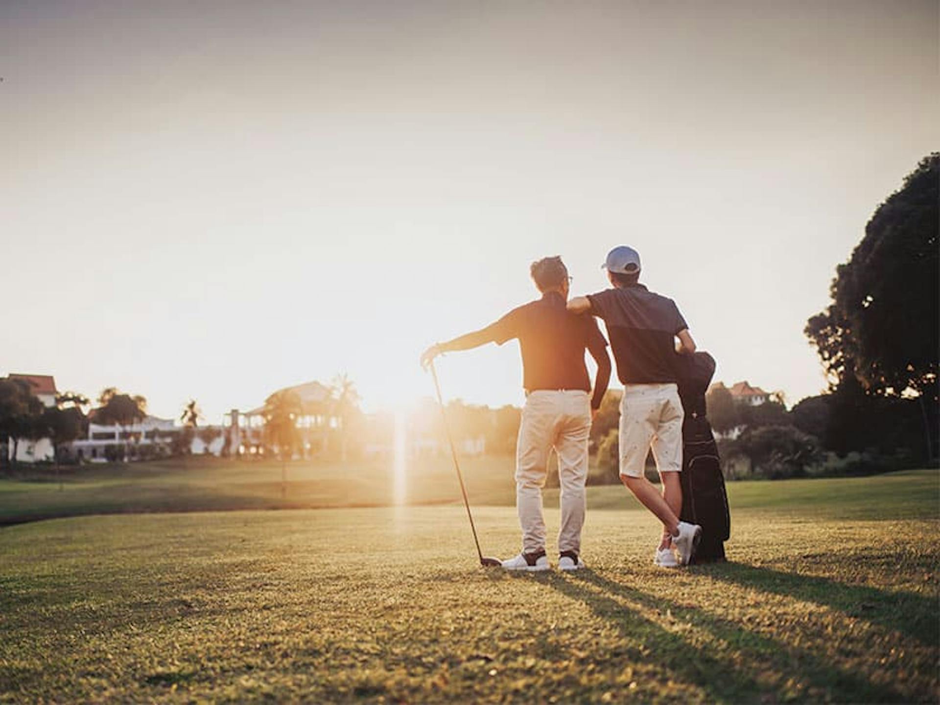 a golfer leans on another taking in the sun that is shining on the horizon