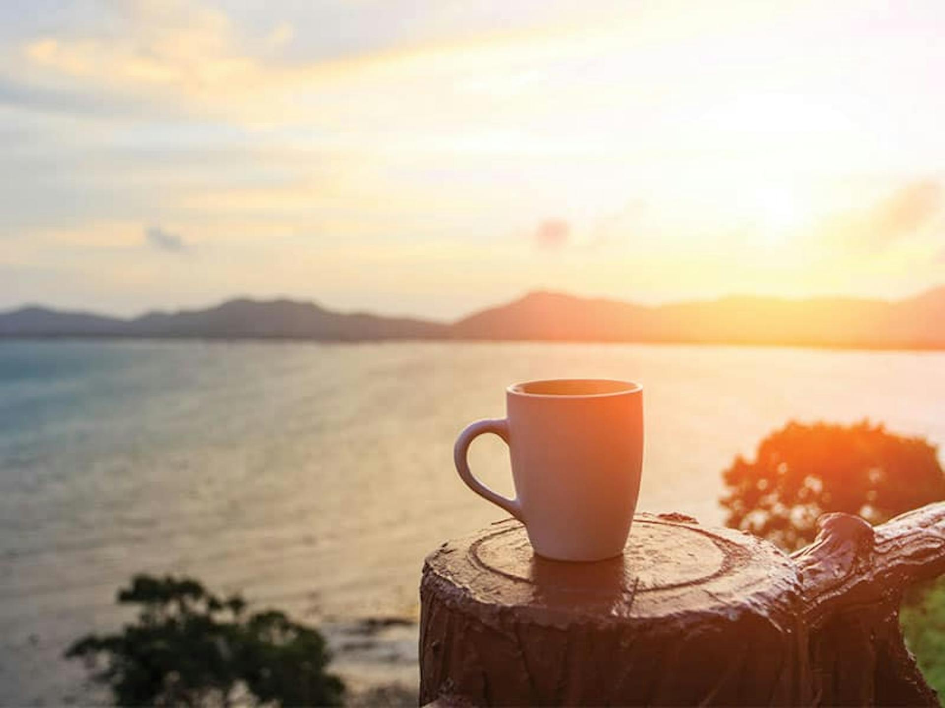 A cup of coffee sits on a wooden fence post with the blurred background of a bay of water and sunset