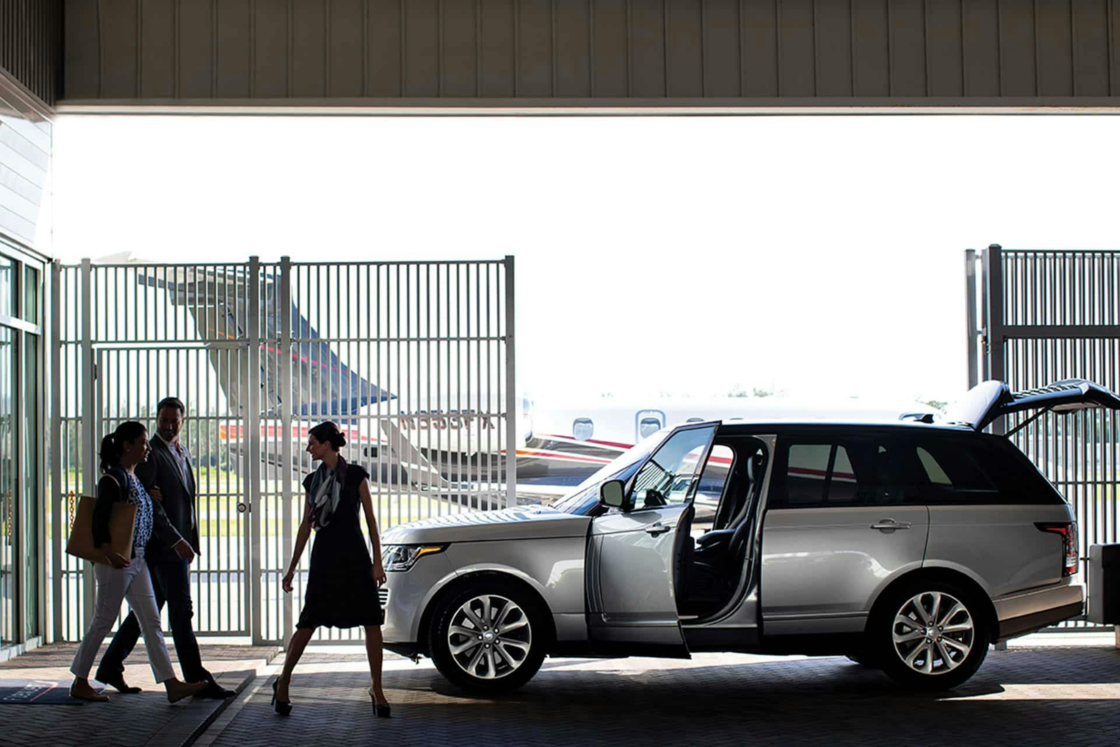 A luxury car waits with open doors outside of a terminal where passengers are walking to be picked up